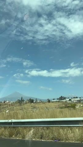 The video was shot from inside a car, providing a view of a field, houses, and a mountain range under a partly cloudy blue sky. In the foreground, the roadside consists of tall, dry grass next to a grey guardrail. Beyond the guardrail, a wide field stretches toward a cluster of houses. Further in the distance, the silhouette of a mountain range dominates the landscape. The peaks are capped with a light covering, possibly snow or ice, enhancing their visual contrast against the sky. The sky itself is vibrant blue, scattered with fluffy white clouds that add depth and texture to the scene. There is the reflection of what appears to be a person in the glass.