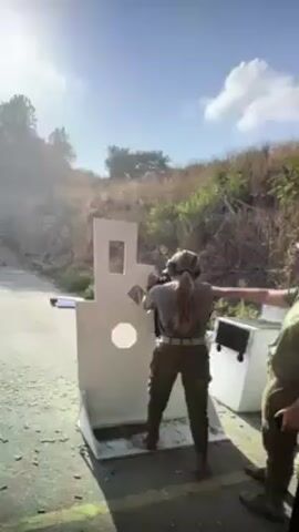 Here is a concise description of the video:

The video shows a woman at an outdoor shooting range. She is wearing a gray t-shirt, green pants, and a belt, and her hair is pulled back in a ponytail. She is standing in front of a white target board with various geometric shapes cut out. She is holding a rifle and shooting at the target. A man in a green shirt and ear protection is standing behind her, likely providing instruction or supervision. The environment consists of a paved area, a grassy hillside with trees, and the partially sunny sky above. The action is centered around the woman firing at the target, causing small white fragments to spray out.