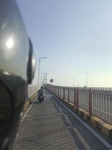 The video opens with a close-up of the left side of a motorcycle helmet, blurry and out of focus. The camera then pans to the right, revealing a two-lane bridge with a white railing on the right and a red and yellow striped barrier on the left. Several streetlights are visible along the bridge, and a speed limit sign indicating "80" is mounted on a pole. Two motorcycles are seen traveling away from the camera on the bridge. The first motorcycle has a rider wearing a white helmet and carrying a large load of what appears to be produce or supplies in bags on the back. The second motorcycle, further ahead, also has a rider. The sky is clear and bright blue, with the sun visible in the distance on the right side of the frame. The bridge extends into the distance, with the sun reflecting on the water below. The video ends with a view of the sun and railing on the right side of the bridge.