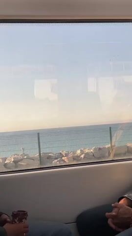 The video shows a view from inside a train, looking out a window at the ocean. A rocky shoreline is visible, separated from the ocean by a fence. The sky is a pale blue and the water is turquoise. The reflection in the window shows parts of the train interior.  Two people are sitting in the foreground, a brown paper cup with a red logo is visible in the hand of the person on the left.  The train appears to be moving, as the view outside the window changes.