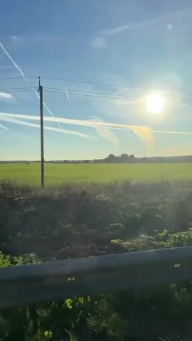 The video shows a view from a moving vehicle looking out at a bright sunny day with a clear blue sky and some white contrails. A green field stretches into the distance with a few trees or buildings on the horizon. In the foreground, there is a roadside barrier and some green vegetation. There are no people visible in the video. The sun is shining brightly and appears to be low in the sky, suggesting it might be morning or late afternoon. The overall impression is one of a scenic countryside drive.