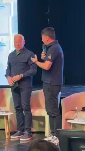 Two men stand on a stage in front of a screen. Both men are dressed in dark blue outfits. The man on the left is fair-skinned with short, white hair. The man on the right holds a microphone. He is speaking and gesturing toward the other man. There are two small white tables with water bottles on them and two pale pink armchairs.
