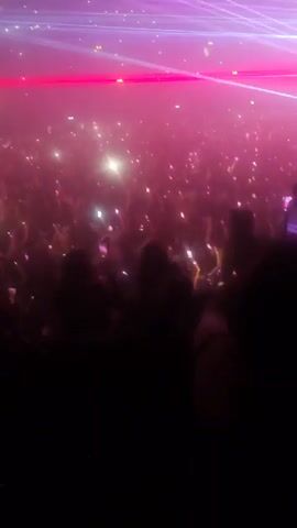 The video captures a densely packed crowd at a concert. Many people are holding up cell phones, presumably to record the performance. The stage lights create a dynamic display with beams of white, blue, and red, enhancing the atmosphere of a live music event. The crowd appears to be enjoying the music and atmosphere.