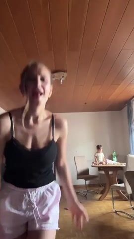A young woman with blonde hair tied back in a bun is dancing in the foreground. She is wearing a black camisole and pink and white checkered shorts. Her arms are flailing and her legs are moving in a rhythmic pattern. In the background, a baby is sitting at a round wooden table, looking towards the camera. The room has wooden paneled walls and ceiling, and a wooden floor. There are chairs around the table, and a doorway is visible on the left side of the frame. The lighting appears to be natural, coming from a window.