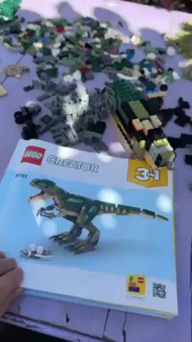 Here’s a description of the video. The camera shows a purple picnic table covered with various Lego pieces. At the center is a Lego instruction booklet for a Lego Creator set, model number 31151, depicting a green and brown dinosaur. Next to the booklet is what appears to be the front section of an in-progress Lego vehicle. A hand briefly touches the instruction booklet. The scene seems to be outdoors on a sunny day.