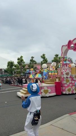 A vibrant parade is captured in this video, featuring a large, ornate float primarily decorated in pink and white with heart-centered white flowers and Hello Kitty motifs. Several characters, including Hello Kitty, and performers in colorful costumes are positioned on the float, some appearing to dance.

In the foreground, a man wearing a blue and white uniform with a matching hat, possibly a park employee or performer, enthusiastically dances and claps along the parade route. Another man in a light blue shirt and khaki pants walks alongside the float, seemingly assisting or monitoring it.

A large crowd of spectators lines the sidewalk in the background, watching the procession. Trees and buildings are visible beyond the crowd. The sky is overcast, indicating a daytime setting. The overall atmosphere appears festive and energetic. The scene is likely taking place at a theme park, possibly Universal Studios Japan given the Hello Kitty theme and location context of Osaka, Japan.