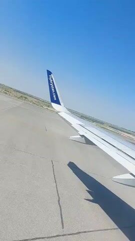A passenger's view from an airplane window shows the wing and the runway. The wing is white with a blue tip featuring the logo "FlyArystan.com". The runway is gray concrete with visible seams. The ground beyond the runway appears dry and sparse, with a clear blue sky above. The plane is on the ground, likely taxiing or preparing for takeoff.