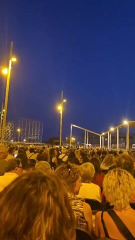 This is a vertical, medium-shot video of a crowd of people gathered outdoors at dusk. The sky is a deep blue, indicating twilight. In the foreground, the backs of several people's heads and shoulders are visible, suggesting the viewer is part of the audience. Their hair colors vary, with blonde and brown tones predominant. One person in the immediate foreground has curly brown hair. Further back, the crowd is dense and extends into the distance. Most individuals are seated, facing away from the camera, towards an area with some structures and lighting.

In the middle ground, a large, open-air structure with a roof supported by pillars is visible. It appears to be a stage or performance area, with lights illuminating its underside. To the left, there are tall lampposts with yellow lights, and a tall, skeletal metal structure that resembles scaffolding or a tower. To the right, more of the covered structure is visible, and beyond that, a person in a dark blue shirt stands facing the crowd. The ground appears to be paved. The overall atmosphere is one of anticipation for an outdoor event, possibly a concert or performance.