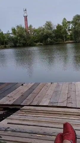 The video shows a first-person perspective from someone resting on a wooden dock with a body of water in front of them. They are wearing red shoes. In the distance, across the water, there is a treeline with a tall, red and white chimney-like structure. The sky is overcast. The water appears calm with ripples.