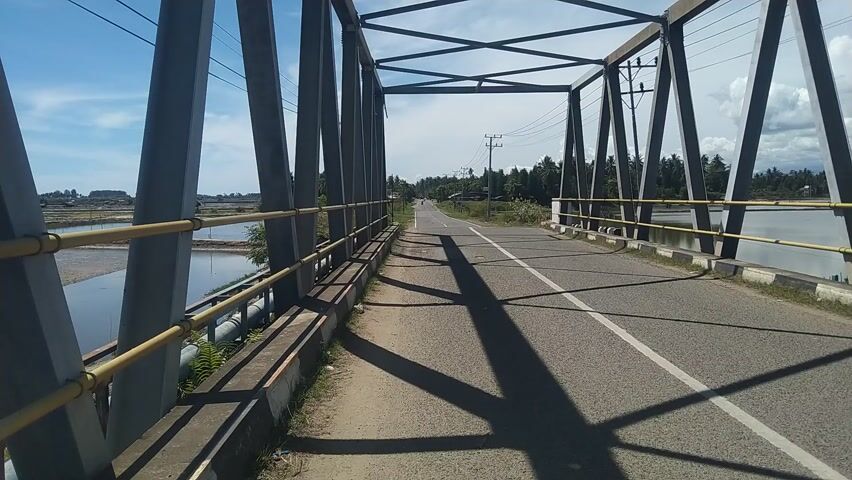 This media file is a video depicting a view from a bridge. The primary subjects are the bridge itself, a road leading over it, and the surrounding landscape. The bridge is a metal truss structure with gray support beams and yellow railings. The road is paved with white lane markings and is casting long shadows, indicating it is likely midday with the sun relatively high in the sky. The weather appears clear and sunny with a blue sky and scattered clouds.

The scene transitions to show the landscape on either side of the bridge. To the left, there are bodies of water, possibly fish farms or rice paddies, with some scattered vegetation and a small hut. The terrain looks flat and rural. In the distance, there are more scattered huts and trees, suggesting a sparsely populated area. The overall setting is a rural area in Indonesia, as indicated by the vegetation, architecture, and landscape.

There are no people visible in the media. The activity taking place is simply the observation of the bridge and its surroundings. Notable details include the contrast between the man-made structure of the bridge and the natural or semi-natural landscape, the clear weather, and the sense of a quiet, possibly warm day. There is no visible text in the media.