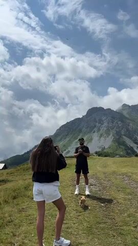 A vertical video shows a person filming with a phone, a man and a dog in the foreground, with a mountain range in the background. The sky is cloudy, and the terrain is grassy. The drone is then released, and the camera pans to reveal a landscape of rolling hills covered in fog, with a wooden cabin in the distance. The drone then flies over the landscape, capturing a panoramic view of the mountains and the valley below. The video ends with a shot of the drone flying towards the horizon.