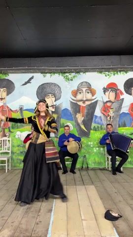 A woman in a traditional black and gold dress dances center stage with her arms moving gracefully. She also wears a small black cap with gold trim and a striped sash. Seated behind her are three men playing musical instruments: one plays a drum, and the other two play accordions. All three men are dressed in blue shirts. They are on a stage with a light wooden floor. Behind them is a mural of cartoon men with distinctive features, set against a background of mountains and a flying bird. A white chair sits to the left of the frame, and a hat lies on the floor to the right.