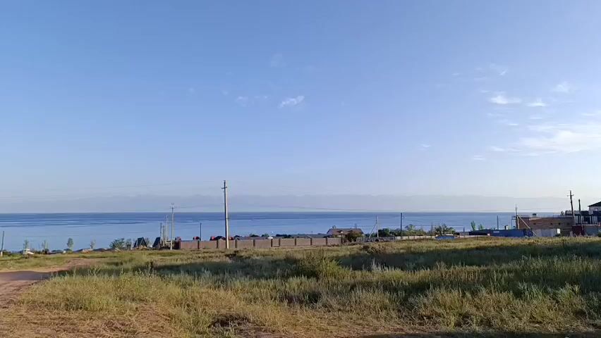 The video shows a wide, dusty dirt road leading towards a body of water, likely a lake or sea, under a clear blue sky with a few scattered clouds. The water is calm and blue, with a distant shoreline featuring hills or mountains. Along the shore, there are several buildings and fences, including what appear to be houses and utility poles. The foreground is dominated by dry, grassy terrain. The camera appears to be mounted on a moving vehicle, slowly traversing the dirt road.