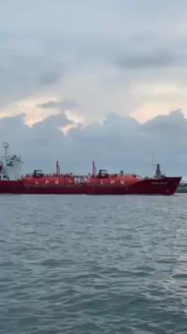 A wide shot shows a red LPG tanker ship moving across the water from left to right. The body of the ship is red and has several cylindrical tanks labeled "LPG" on its deck. The ship has a tall mast and various other structures. The name of the ship "ASIAN GAS 7" is written on the stern. The water is dark and slightly choppy. The sky is overcast with clouds and some hints of a sunset. The camera is stationary, capturing the ship moving across the horizon.