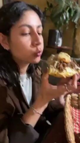 A woman with dark curly hair and a brown jacket takes a bite of food in a cafe. She has multiple earrings and rings on her hands. The food appears to be a large, thick slice, possibly of pizza or flatbread, with brown toppings and a lighter sauce. Behind her, there are plants and what seems to be other seating areas. The lighting is dim and warm.