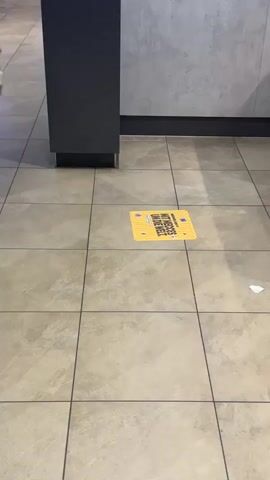The video shows a floor with square tiles. There is a yellow square sign on the floor with black text and a yellow border. A black pillar is on the left side of the frame. In the background, a person wearing blue jeans and a blue shirt is walking away from the camera, carrying a brown paper bag. Another person is visible further in the background.