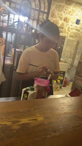 A person is standing behind a counter and holding a stamp and an ink pad. The person is wearing a light-colored t-shirt and has their hair pulled back. There is a pink cup with a straw on the counter in front of the person. Behind the person, there are shelves with various items, including bottles and boxes. The environment appears to be a shop or restaurant.