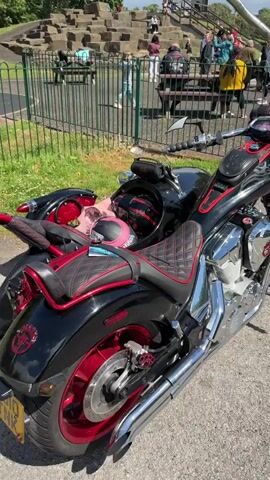 The video shows a close-up, panning view of a customized black and red motorcycle with a sidecar. The motorcycle is parked on a paved surface, likely a path. In the background, a playground with green and silver structures and a slide is visible. Several people are scattered around the playground area, some walking and others standing. The overall environment appears to be an outdoor park on a sunny day. The focus remains on the motorcycle throughout the video.