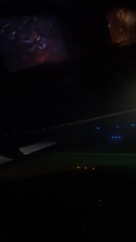 The video shows a view from an airplane window at night. The wing of the plane is visible in the foreground, with lights on the runway below. The lights appear to be blue and yellow, indicating a runway at an airport. There are also some screens visible in the upper part of the video, showing images that are not clear. The overall scene suggests the plane is either landing or taking off at night.