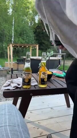 The video shows a person in a striped pajama-like pants, sitting on a wooden deck with a small wooden table in front of them. On the table are two bottles of Corona Extra beer, a white towel, and a small bowl of chips. The person is holding a box with writing and looking at it. Behind the table, there's a backyard area with a small wooden gazebo decorated with string lights, a grill, and a hot tub. The ground is covered with grass and gravel. In the background, there's a line of trees, and the sun is shining through them.