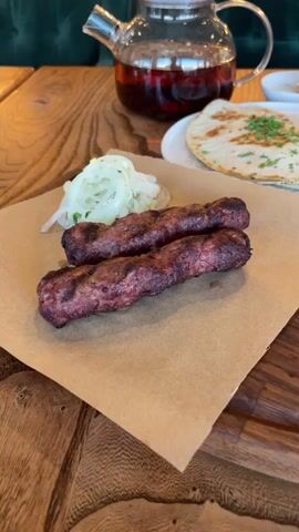 The video opens with a close-up shot of two grilled meat skewers on a brown paper lining on a wooden table. To the left of the skewers is a pile of thinly sliced white onions and chopped green herbs. Behind the skewers, on a white plate, is a folded flatbread topped with chopped green herbs. To the right of the flatbread is a small white ramekin of sauce. In the background, a glass teapot filled with a dark liquid, presumably tea, sits on a dark wooden surface. Further back, a slice of layered cake with a light brown frosting is visible on a white plate, next to a small white bowl. The environment appears to be a restaurant setting, with a green upholstered seat visible in the upper left corner. The camera pans slightly to the right, focusing more on the flatbread and the sauce, then to the cake.
