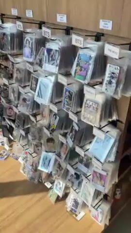 The video shows a wall display of packaged photocards, sorted by K-pop group such as BTS, TXT, and ENHYPEN, among others. Above each group's section is a label with the group's name. The individual holding the camera reaches for a BTS photocard, removes it from the display, and shows it to the camera. Signs at the top of the display state that the photocards are "photocard playpad" and "photocard papel," with prices of R$32.00 and R$58.00, respectively. The display is mounted on a wooden wall with a wooden floor visible below.