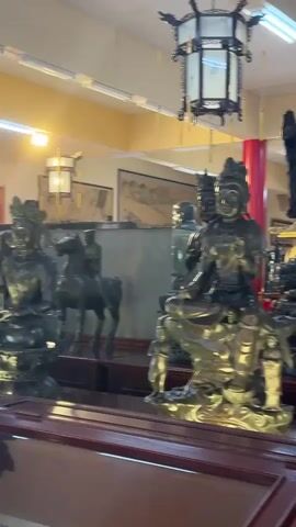 The video pans across a display of various bronze statues, likely in a temple or shrine. The statues appear to be of religious or cultural figures, with some depicting seated individuals, others standing, and one on horseback. The lighting suggests an indoor environment, possibly with fluorescent lights overhead and some decorative lanterns. The background shows more statues, red columns, and what appear to be paintings or scrolls on the wall. The camera moves smoothly, offering a clear view of the detailed craftsmanship of the statues.