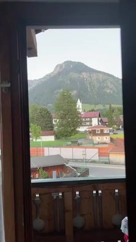 The video shows a view from an indoor balcony, looking out over a small European town nestled in the mountains. A mountain dominates the background, covered in green trees. Below it is a cluster of buildings, including a church with a white spire. There is a tennis court, a street with cars, and various wooden structures visible. The balcony railing is made of wood with decorative cutouts. The sky is overcast.