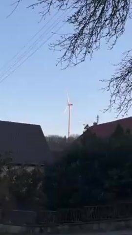 The video shows a view of a rural or suburban landscape, featuring several wind turbines in the background against a clear blue sky. In the foreground, there are houses with dark-colored roofs and dense green bushes partially obscuring the view. Some tree branches are visible in the upper corners of the frame, adding a natural element to the scene. The wind turbines appear tall and modern, contrasting with the more traditional architectural style of the houses. Electrical power lines are strung across the sky. The overall impression is one of a peaceful setting where renewable energy sources are integrated into the local environment.