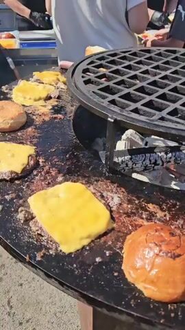 A video shows a circular grill with a grate on top. Several cheeseburgers are cooking on the flat surface of the grill. Some of the cheeseburgers have melted cheese on top, while others have buns. The grill is heated by glowing embers underneath. In the background, people are visible preparing food. One person in a black apron and black gloves is using a metal grater over the grill. Another person in a black t-shirt and apron is smiling. A third person in a red t-shirt and apron is also visible. The setting appears to be an outdoor food event or festival.