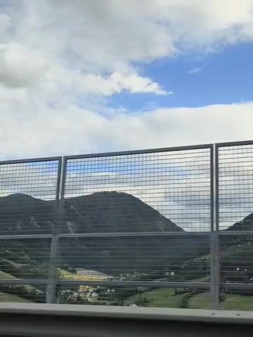 The video shows a view through a metal fence of mountains and a cloudy sky. The fence is made of interconnected metal squares. In the distance, there are green hills and a valley with some buildings. The sky is blue with scattered white clouds. The perspective suggests the video is being filmed from a moving vehicle, possibly on a highway.