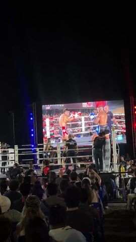 The video depicts a live Muay Thai boxing event, showing a crowd of people watching a boxing match on a large screen. The screen displays a live feed of two boxers in a ring, engaged in combat, with referees present. The boxing ring itself is set up within the venue, though the main action is viewed via the screen. The audience is diverse, with people of various ages and ethnicities. Some are sitting in chairs, while others are standing, and many are holding up their phones, likely to capture the moment. The event appears to be held outdoors at night, indicated by the dark background and the presence of stage lighting, including spotlights and colored lights.