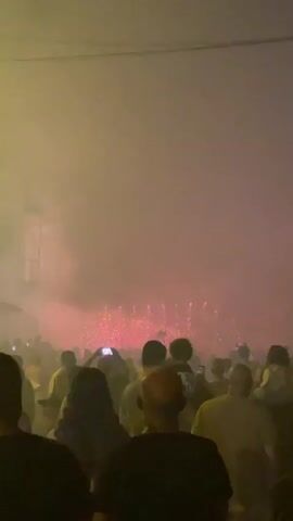 A crowd of people are watching a fireworks display. The fireworks are red and pink and are exploding in the distance. There is a lot of smoke in the air, which is illuminated by the fireworks. The crowd is facing away from the camera, towards the fireworks. Some people are holding up their phones to record the display. The scene appears to be at night.