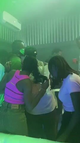 A group of people are dancing in a nightclub. The lighting is a mix of purple, blue, and green, creating a vibrant atmosphere. The individuals are dressed casually, with one person wearing a bright pink tank top and another in a white t-shirt. The camera focuses on a cluster of people in the foreground, with others visible in the background. Some individuals appear to be interacting closely, possibly dancing together. The environment suggests a lively and energetic social gathering.