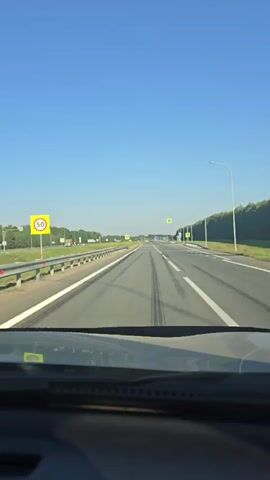 A first-person perspective video shows a clear, sunny day with a bright blue sky as a car travels down an empty highway. The road is paved with asphalt and marked with white lines. There are guardrails on the left side of the road and trees lining the right side. Pedestrian crossing signs are visible on the right side of the road. The dashboard of the car is visible in the foreground. There are no other vehicles or people visible in the video.