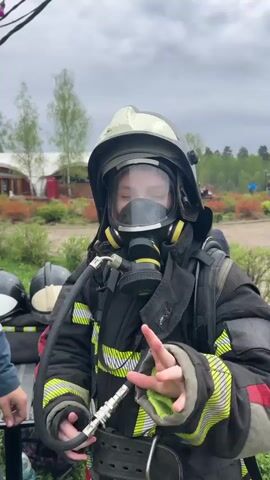 A child dressed as a firefighter stands facing the camera. They are wearing a black and yellow fire suit, a helmet with a visor, and a gas mask. They are connected to an air tank on their back via a hose. The child appears to be on a set, with a table to their left holding other helmets and gear. There are buildings, trees, and other people visible in the background. The sky is cloudy.