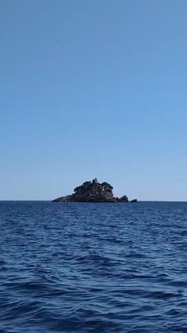 The video shows a small, rocky island in the middle of a vast, blue ocean under a clear sky. The island is covered with trees and appears to have a small structure on top, possibly a lighthouse or a building. The water is choppy, with visible waves. The camera is moving, suggesting it is being filmed from a boat. The overall impression is one of isolation and tranquility.