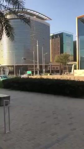 The video shows a cityscape on a sunny day. In the foreground is a pedestrian area with light gray benches and stylized metal railings. To the left is a palm tree. Across the street are various buildings, including a large cylindrical building with a flat roof, a brown building, and a building with turquoise accents. There are streetlights and a few vehicles on the road. The sky is clear and blue.