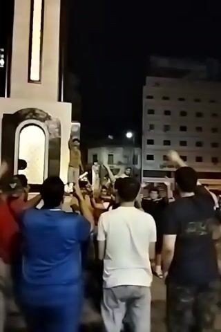 At night, a crowd of men, mostly young, gather at the base of a tall, lit monument or sculpture. Some of the men are recording videos with their phones of a man who has climbed onto the monument and is waving his arms. The building looks modern with glass and white paneling. Behind the group, there are more buildings with visible windows and street lights. The atmosphere seems festive.