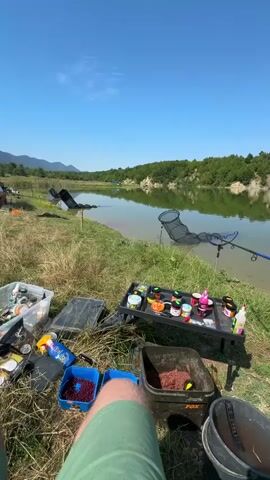 The video shows a fishing setup on the bank of a lake or pond. The foreground is dominated by fishing gear, including a tackle box filled with various containers, a blue container with red bait, and a larger bucket with what appears to be groundbait. A small black table holds an assortment of jars and bottles, likely containing fishing attractants or bait additives. Two fishing rods are set up, one with a reel and a landing net extended over the water. A person's legs, wearing green shorts, are visible in the lower center of the frame, suggesting they are seated. The background features a calm body of water reflecting a clear blue sky and a tree-lined shore with a sandy bank. Several other fishing rods and nets are visible further down the bank. The overall impression is a serene outdoor setting for a day of fishing.
