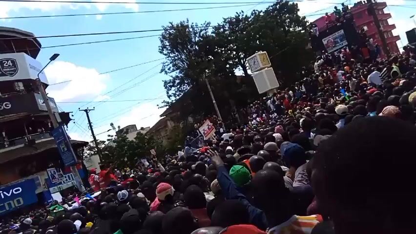 Here is a detailed description of the video:

The video shows a large crowd of people gathered in an urban area. The crowd appears to be mostly composed of black individuals, many wearing casual clothing and hats of various colors. The crowd is densely packed, with people standing shoulder to shoulder. In the background, there's a building with advertising on its side. On the right side of the crowd, there’s a raised platform on top of a building, where more people are gathered. Above the crowd, there is a large white box hanging from wires. The sky is visible with a light blue color and scattered clouds. The camera moves a bit, but mainly keeps a wide shot of the crowd.