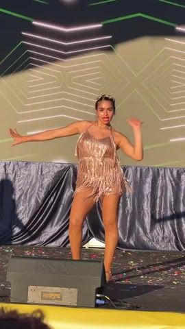 A female dancer performs on a stage, wearing a sparkly, fringed dress and a jeweled headband. The stage is decorated with confetti and draped with dark fabric. A large screen in the background displays geometric green and white light patterns. The dancer moves dynamically, with her arms and legs extended in various poses. The lighting is bright, suggesting an outdoor daytime performance.