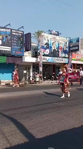 A street parade is taking place under a clear blue sky. Two individuals in traditional costumes, one in red and gold with an elaborate headdress holding a framed portrait, and another in a white outfit with decorative sash and headwear, are walking down the street. Following them is a banner that appears to read "INDONESIA 81". A procession of people in white uniforms, some carrying flags, marches behind the banner. On the right side of the street, a crowd of spectators, mostly women wearing hijabs, are sitting on the ground watching the parade. The background features buildings with advertisements and some parked motorcycles. The sun is casting shadows on the road.