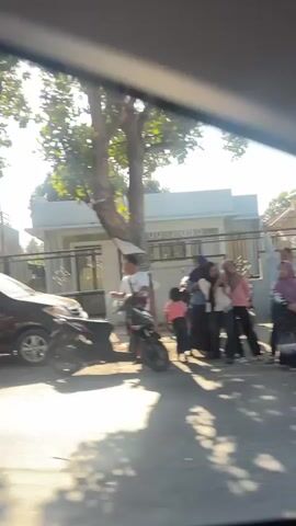 A brown SUV is parked on the right side of a road. A man wearing a white shirt and black pants is standing next to a black motorcycle. The man is holding a cigarette in his right hand and is looking to his right. Behind the man, a group of people in white uniforms with red sashes and crowns are gathered on the side of the road. The people appear to be a marching band or a performance group. A yellow Mitsubishi van is parked on the left side of the road. The video shows a street scene with houses, trees, and buildings in the background. The sun is shining and the sky is clear. The overall atmosphere is one of a community event or parade.