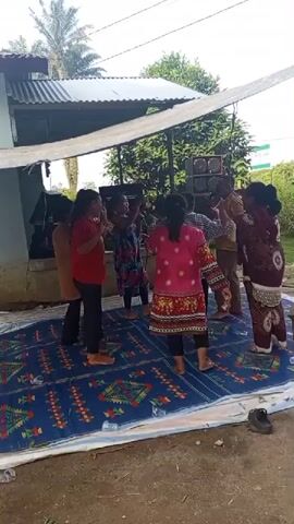 A group of women are dancing on a blue patterned rug outdoors. They are under a makeshift canopy supported by poles. In the background, there is a house with a corrugated metal roof and some speakers. The women are wearing colorful clothing and appear to be enjoying themselves. The ground around the rug is dirt and some debris.