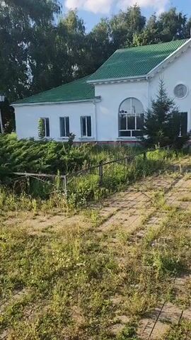 The video shows a white building with a green roof and arched windows. A path made of paving stones leads up to the building, which is overgrown with grass and weeds. There are trees and bushes around the building. The sky is blue and clear.