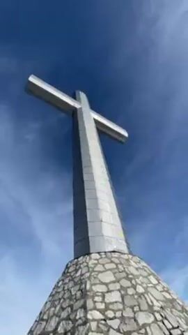 The video starts with a low-angle shot of a large, silver cross standing on a stone base against a clear blue sky with some wispy clouds. The camera then pans down to show a group of people standing on a snowy mountaintop, near a metal railing. A young man in a white tank top stands out in the foreground, looking at his phone. The camera then pans across a breathtaking view of a sea of clouds stretching out to the horizon, with the sun shining brightly through the sky. The video ends with a view of the cloudscape and some vegetation in the foreground.