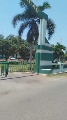The video opens with a view of an archway with green and white stripes. The words "ITBANGAD" are visible on the arch. Beyond the archway, a paved road leads into a grassy area with trees and a building. A green metal gate is partially open. The camera pans to the right, revealing a white fence with decorative pillars and more trees. Further on, a building with a dark tiled roof is visible, along with a speed limit sign indicating "MAX 10 KM". The scene appears to be an entrance to a facility or compound, possibly military or governmental, given the signage and the overall orderly appearance. The sky is clear and blue, suggesting a sunny day. There are no people visible in the video.