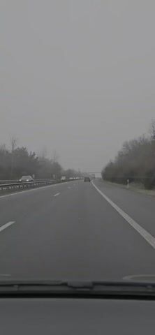 The video shows a gray highway view from the windshield of a car, with cars traveling ahead. Bare trees flank the road, and an overpass appears in the distance. The sky is overcast, contributing to the overall grayness of the scene.