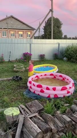 A toddler wearing a pink t-shirt and yellow shorts stands in a grassy yard near two inflatable pools. The larger pool, decorated with watermelons, is filled with murky water, while the smaller, yellow pool contains clear blue water. In the background, a light-colored house with visible windows stands behind a corrugated metal fence. A bicycle and a scooter are visible near the fence, along with some scattered items. The sky above is a gradient of pink and white clouds, suggesting either dawn or dusk. The scene appears to be a domestic outdoor setting.